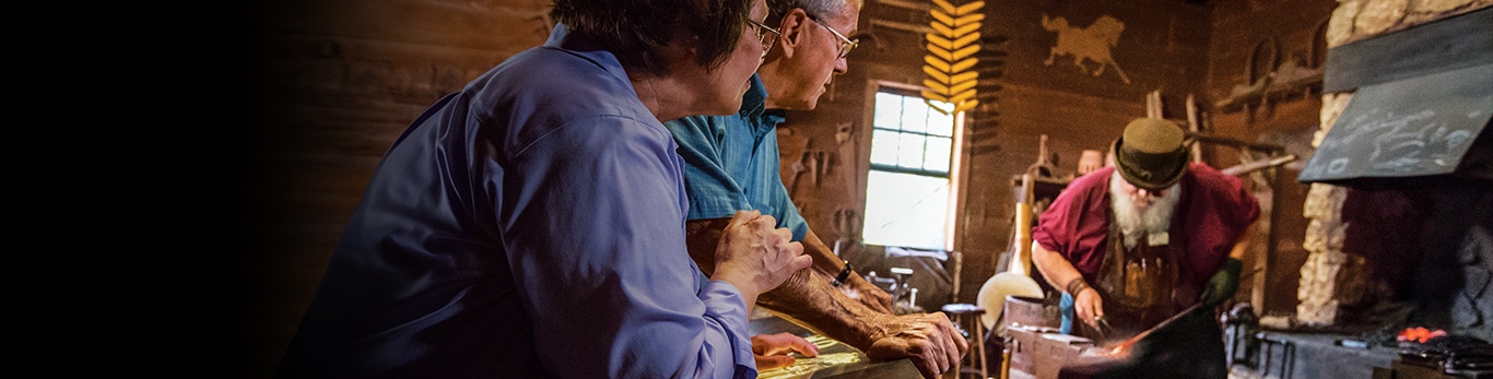 Vyresnio amžiaus pora stebi kalvio gaminamą metalinio plaktuko dalį istorinėje vietoje Grand Detour, Ilinojuje (Jungtinėse Valstijose)