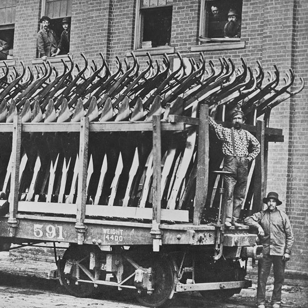 1882 m. nuotrauka – trys vyrai stovi šalia Deere & Co traukinio vagono, prikrauto plieninių plūgų ir paruošto išgabenti, o fabriko darbininkai žūri pro langus iš užnugaryje esančio pastato
