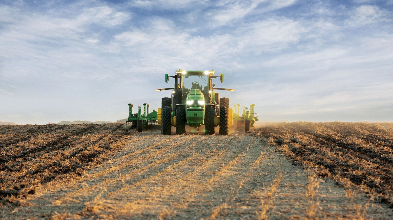 Savaeigis John Deere traktorius, traukiantis žemės dirbimo įrangą per atvirą lauką.