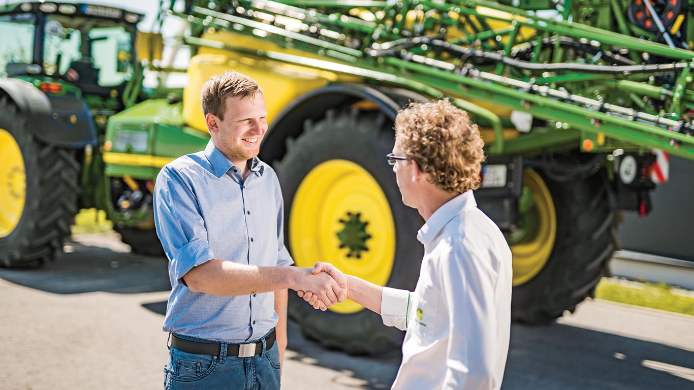 Rankų paspaudimų pardavėjas ir klientas priešais John Deere purkštuvą