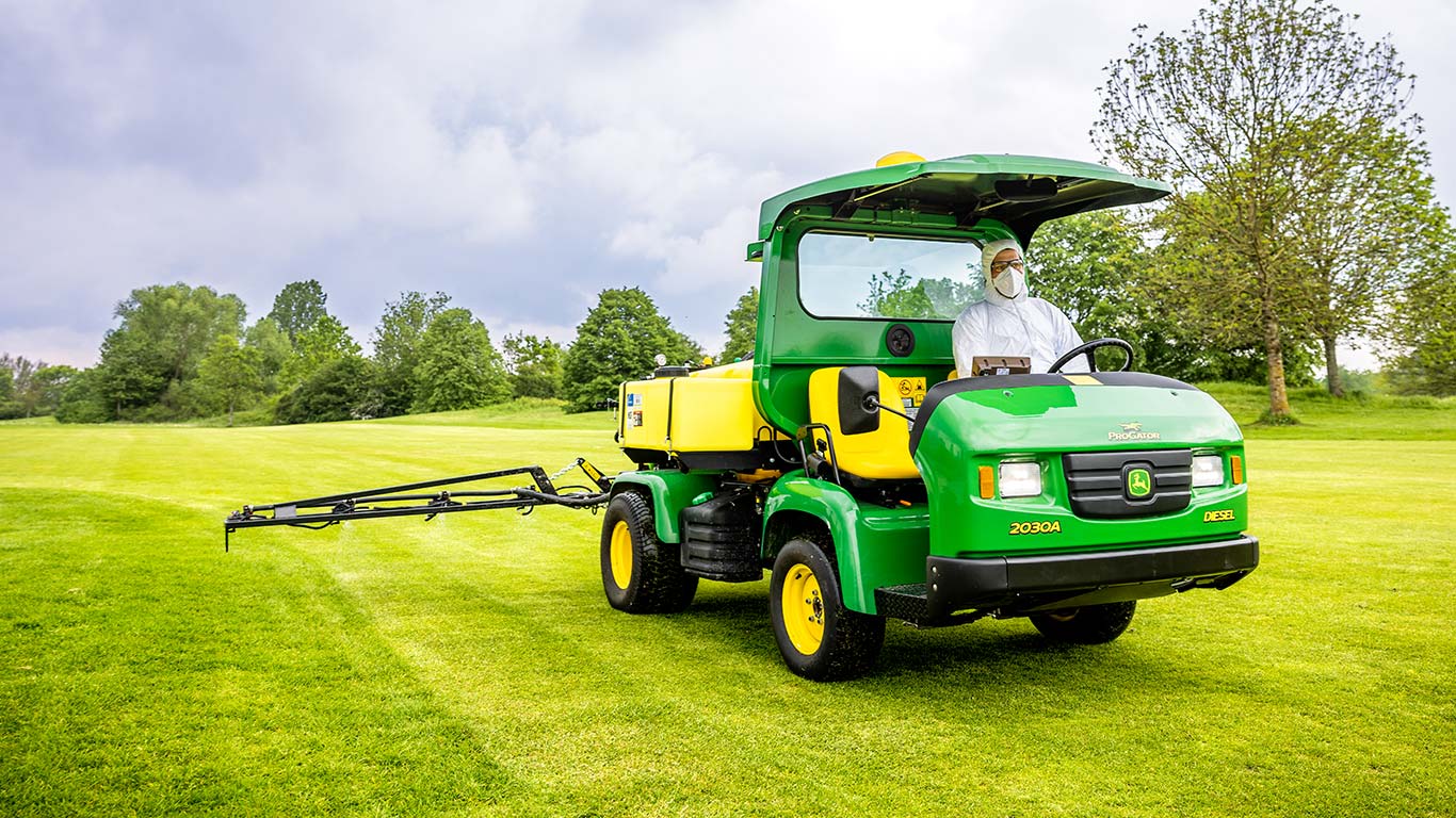 Vejos prižiūrėtojas, vilkintis apsauginiais drabužiais, purškia golfo aikštyno veją John Deere purkštuvu.
