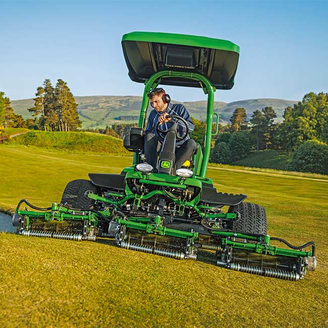 Iš priekio matomas vejos prižiūrėtojas, dirbantis vejos plote su John Deere užsėjamąja aikštynų vejapjove.