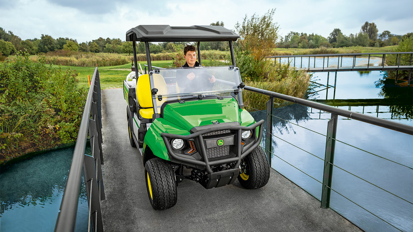 John Deere Gator GS universalusis visureigis važiuoja per siaurą tiltą nebanguojančiu vandens keliu, apsuptu vešlios žalumos ir medžių. John Deere Gator GS universalusis visureigis važiuoja per siaurą tiltą nebanguojančiu vandens keliu, apsuptu vešlios žalumos ir medžių.