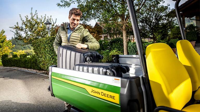 Asmuo žalumos ir medžių apsuptyje, pakraunantis didelį juodą lagaminą į "John Deere Gator GS" krovinių skyrių su ryškiai geltonomis sėdynėmis.