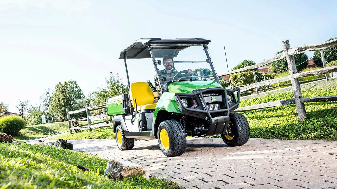 Žalias "John Deere Gator GS" universalusis visureigis važiuoja asfaltuotu keliu per apželdintą teritoriją su žole ir medinėmis tvoromis.