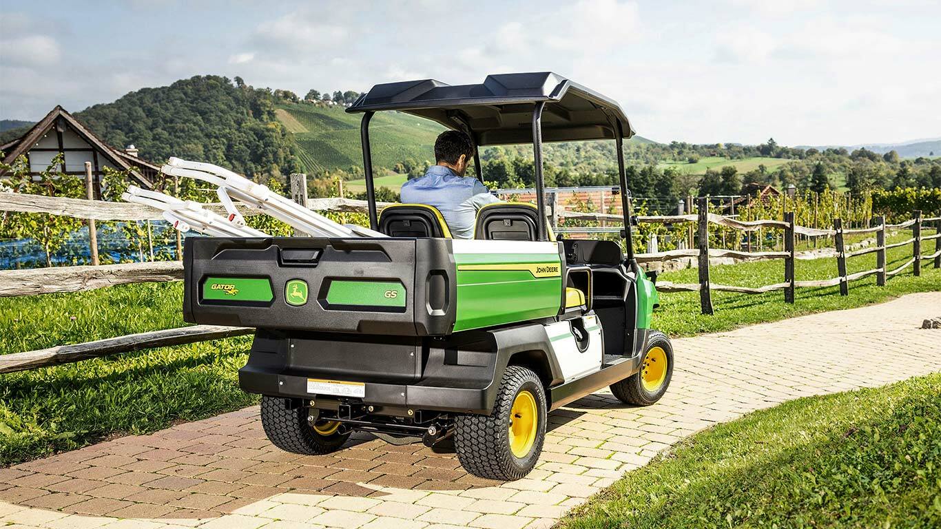 "John Deere Gator GS" vaizdas iš galo važiuojant plytelėmis grįstu keliu su vaizdingomis kalvomis ir vynuogynais fone.
