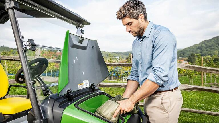 Asmuo, įstatantis paminkštintą maišą į priekinį žaliojo "John Deere Gator GS" daiktadėžę, pastatytą lauke šalia medinės tvoros ir vynuogyno.