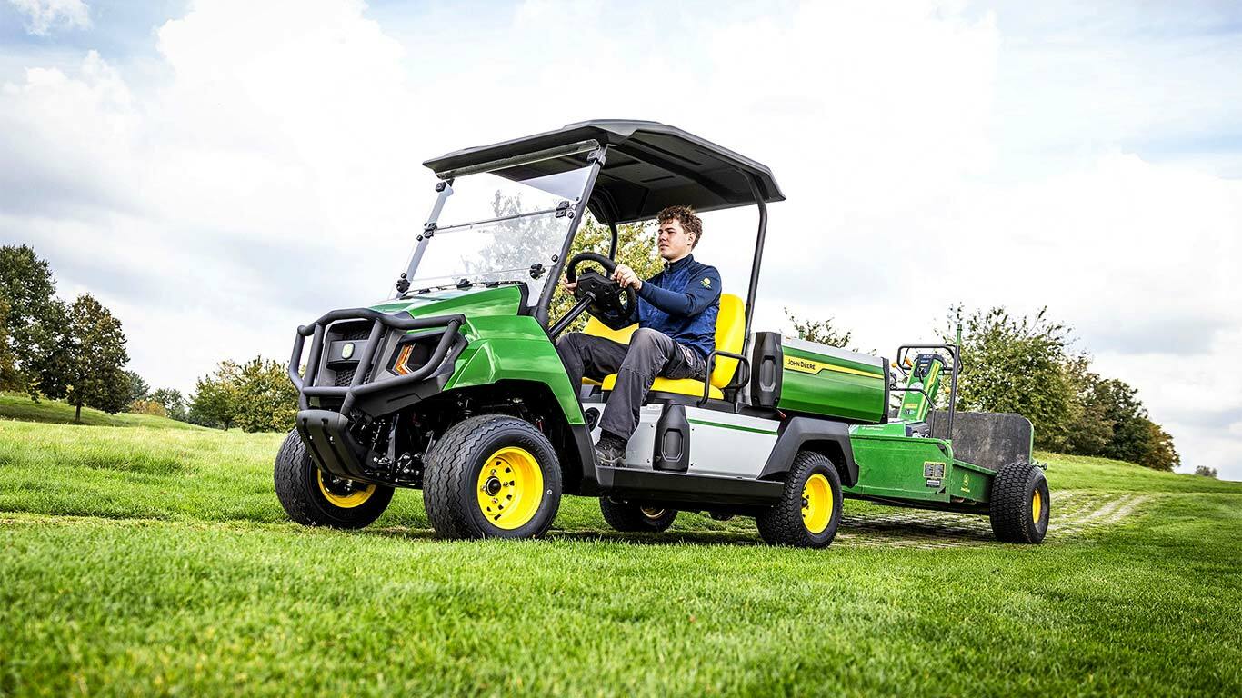 Žaliojo "John Deere Gator" elektrinio universaliojo visureigio vilkimo įranga pievoje po šiek tiek debesuotu dangumi.