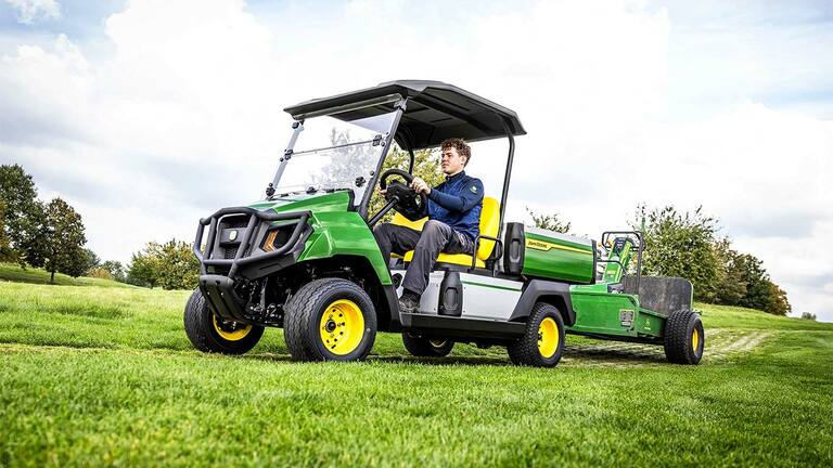 Žaliojo "John Deere Gator" elektrinio universaliojo visureigio vilkimo įranga pievoje po šiek tiek debesuotu dangumi.