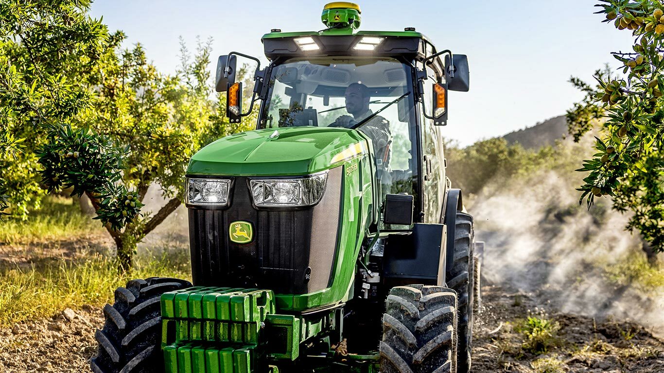 Žalias ir geltonas „John Deere“ traktorius 5075EN važiuoja sodo gruntiniu keliu, apsuptas vaismedžių ir saulės šviesos.