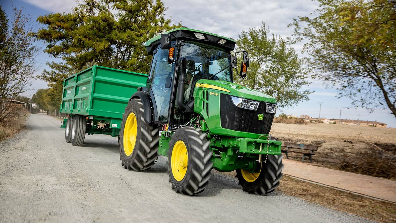 John Deere 5075EN traktorius tempia žalią priekabą kaimo keliu su medžiais fone.