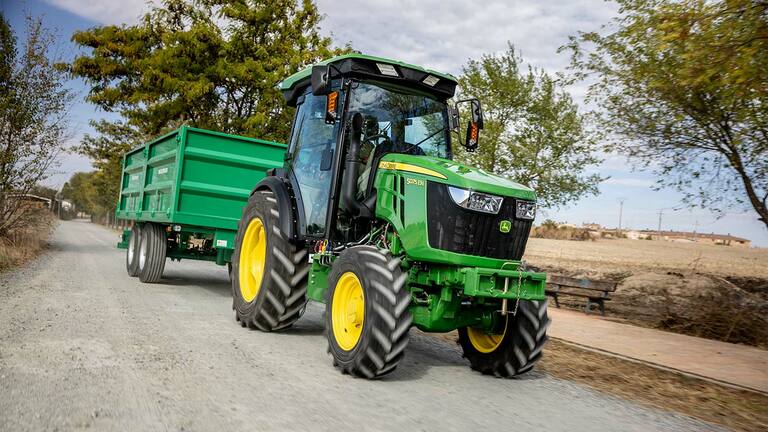 John Deere 5075EN traktorius tempia žalią priekabą kaimo keliu su medžiais fone.