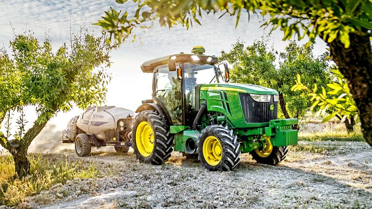 Žalias ir geltonas „John Deere“ traktorius 5090EN, saulėtą dieną traukiantis purkštuvą vaismedžių sode.