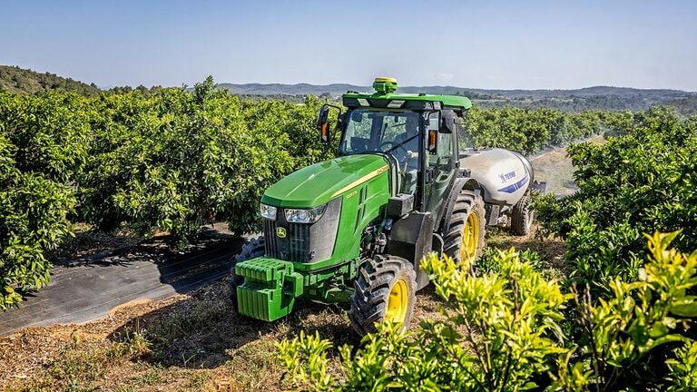 Žalias ir geltonas „John Deere“ traktorius 5105EN velka vešliame sode su fone matomomis kalvomis.