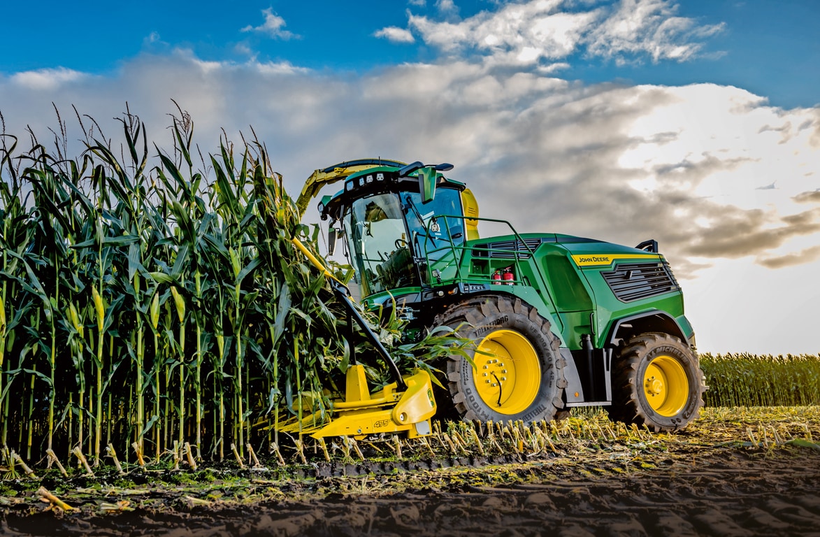 "John Deere" pašarų kombainas, imantis kukurūzų derlių lauke po mėlynu dangumi su debesimis.