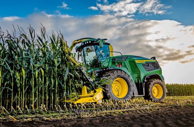 "John Deere" pašarų kombainas, imantis kukurūzų derlių lauke po mėlynu dangumi su debesimis.