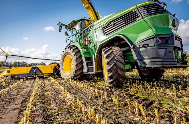 "John Deere" pašarų kombainas dirba nuimto derliaus kukurūzų lauke, o jo didelės padangos ir pjovimo mechanizmas matomi giedro dangaus fone.