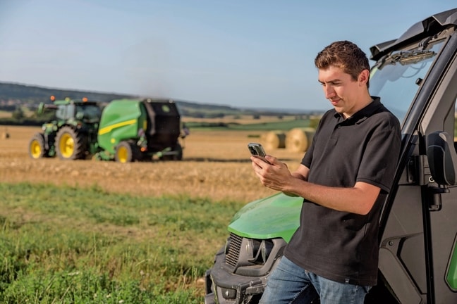 Žmogus mobiliajame telefone tikrina ryšulių dokumentų duomenis, fone matomas "John Deere" VR ryšulių presas